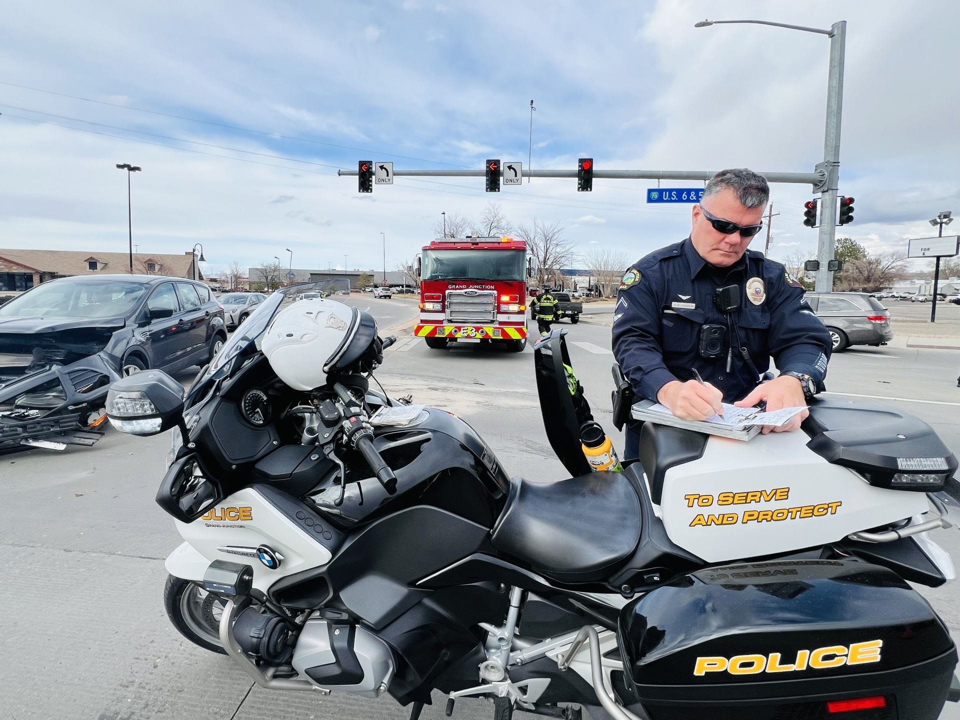 Photo of GJPD Motor Officer at his motorcycle in an intersection. Firetruck in background.